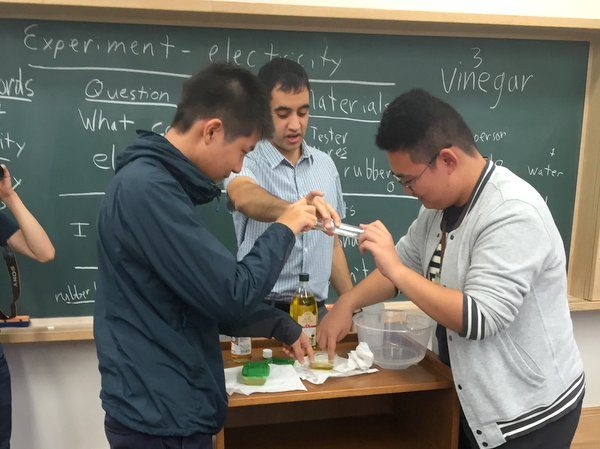 Experiment - electricity と書かれた黒板の前で、外国人講師と男子中学生2人が実験している
