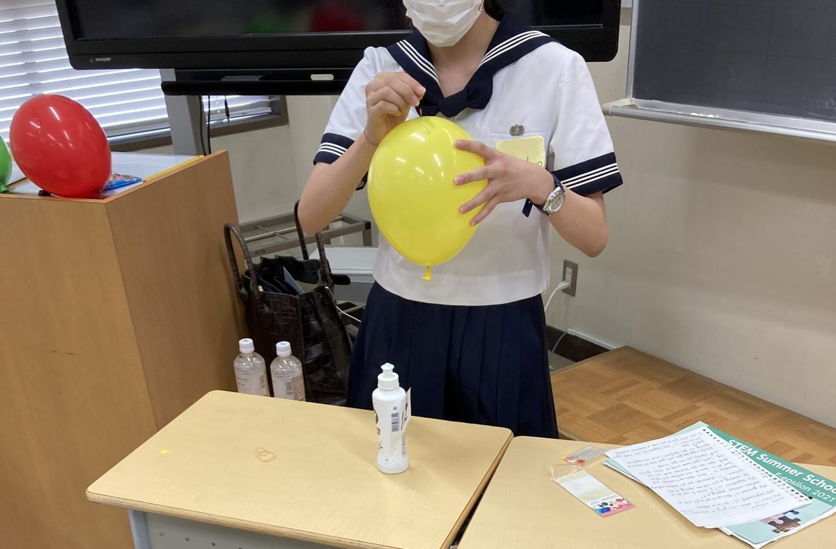 教室で黄色い風船を膨らませる実験に取り組むセーラー服の中学生