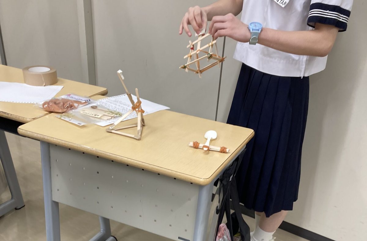 木の棒と輪ゴムで組んだ構造物を手に実験する中学生