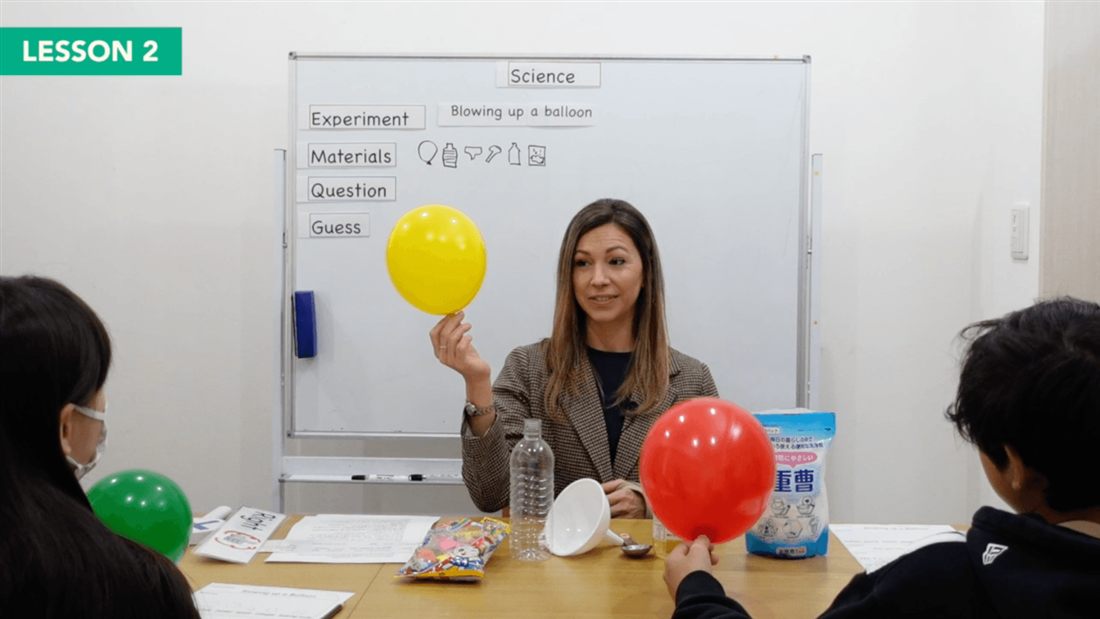 Experiment・Materials・Question・Guessのカードが貼られたホワイトボードの前で、風船を使って授業をする先生と子どもたち